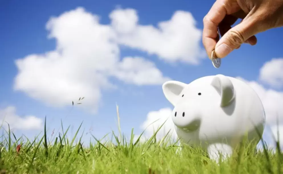 Mano-metiendo-una-moneda-en-una-hucha-de-cerdito-en-un-campo-con-un-cielo-azul
